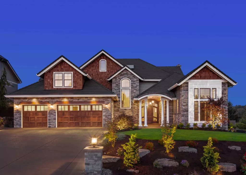 Wood finish garage doors with small windows across the top on a large brick-and-stone home at dusk.