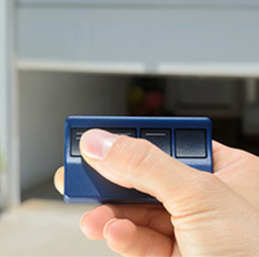 Person's hand pressing a button on a small blue garage door opener remote. Open garage door in background.