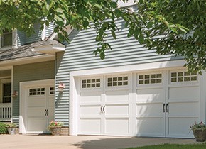 White double-garage doors with small windows at the top, on attached garage of house with light blue siding.